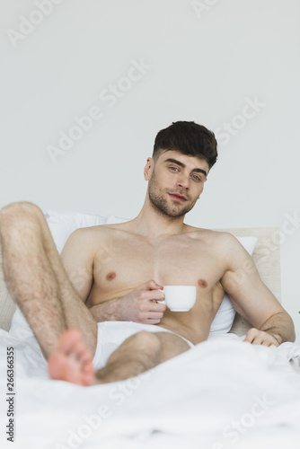 selective focus of good-looking young man in underwear lying in bed with coffee cup and looking at camera