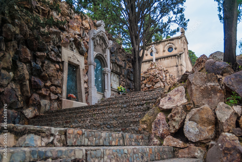 Low-angle view of the stairs and the stone wall with graves and crypts on the Montjuic Cemetery in overcast day, Barcelona, Catalonia, Spain
