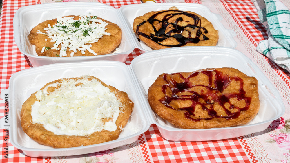 Langos, typical Hungarian food specialty, with sour cream, cheese, dill, chocolate syrup, jam on plastic plates for take away on a table, four deep fried donuts in direct sunlight at a food market Langos, typical Hungarian food specialty, with sour cream, cheese, dill, chocolate syrup, jam on plastic plates for take away on a table, four deep fried donuts in direct sunlight at a food market