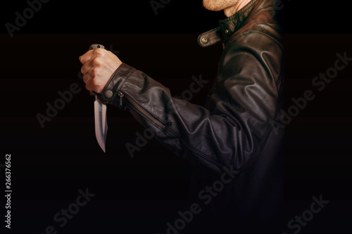 A man in a black leather jacket with a knife in his hand on black background