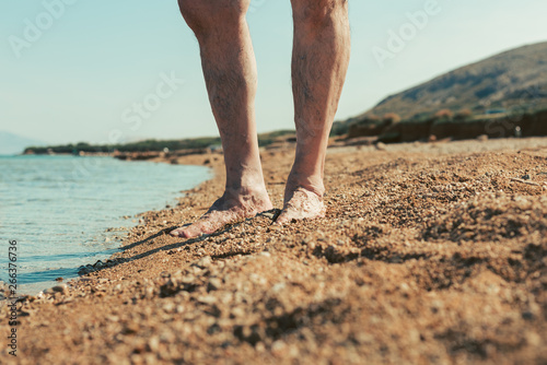 Close up of male hairy legs on yellow sandy beach with sea and hill on background. Male footsteps in the sand. Man standing by the beach. Vacation and Travel concept. Enjoying the summer and sea. © Matt Benzero