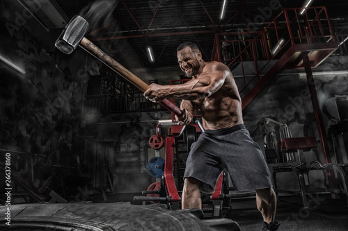 Sport Fitness Man Hitting Wheel Tire With Hammer Sledge Training, Young Healthy Guy Gym Interior photo