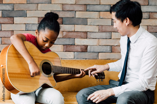 Young Asian male teacher giving a guitar lesson and teaching how to play guitar to African American girl in indoors room photo