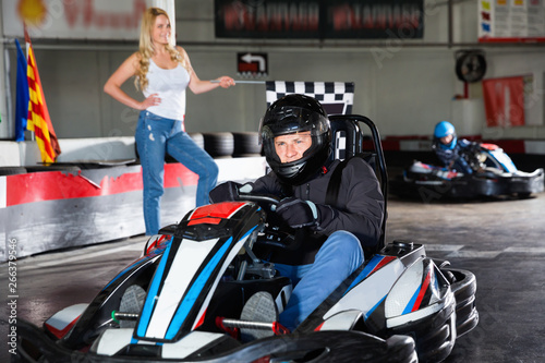 Man in helmet driving car for karting in sport club, woman with flag on background © JackF