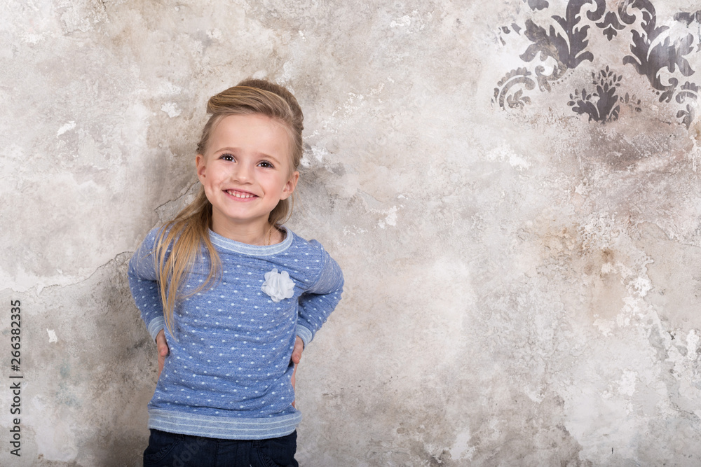Portrait of a little attractive smiling girl in a blue sweater and pants with hair folded into her hair against a grunge wall background. Portrait of a little attractive smiling girl in a blue sweater and pants with hair folded into her hair against a grunge wall background.