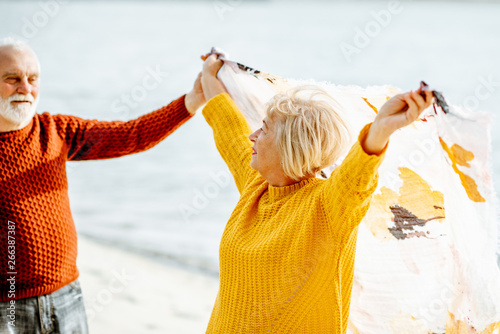 Lovely senior couple playing with scarf during the windy weather, standing together on the sandy beach, enjoying free time during retirement near the sea #266387387