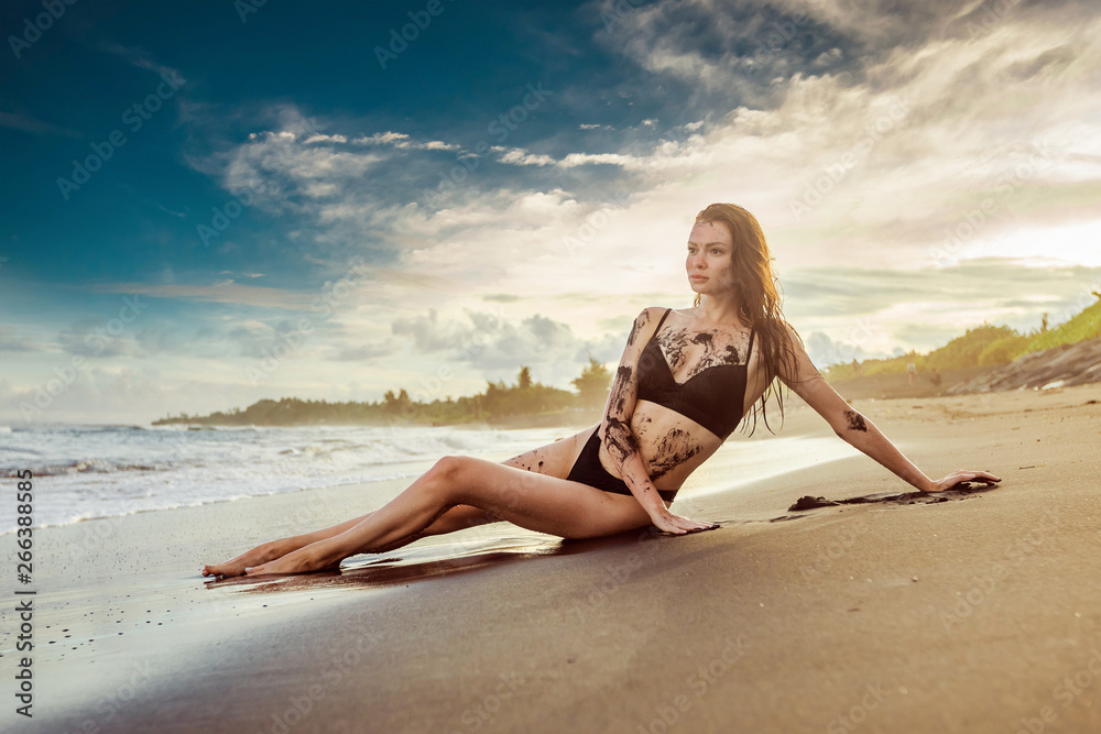 The girl in a bathing suit covered with black sand is lying on the beach. In the background sunset and the ocean