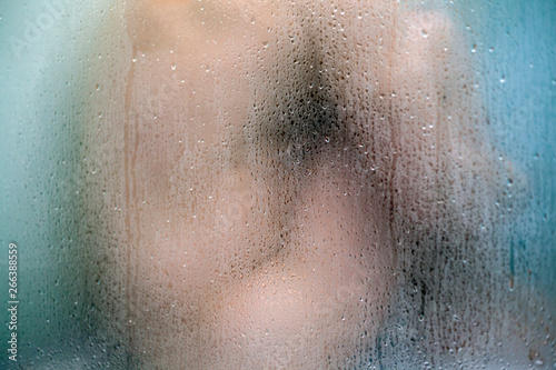 A nude man is seating in the shower bath. Hea blurred silhouette is visible through the sweat glass surface with streams and water drops.