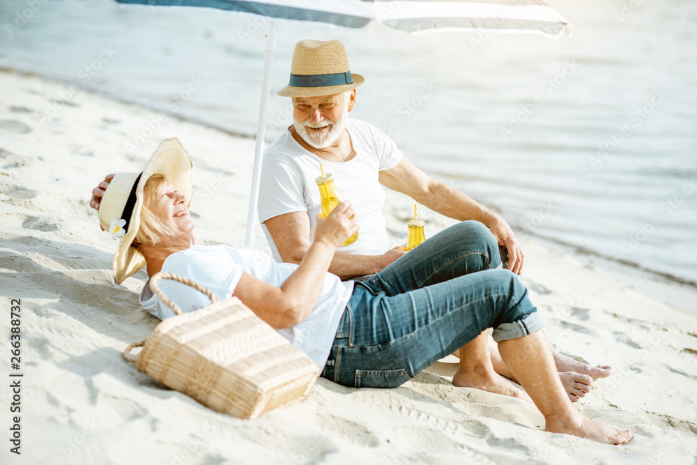 Happy senior couple relaxing, lying with drinks on the sandy beach, enjoying their retirement near the sea