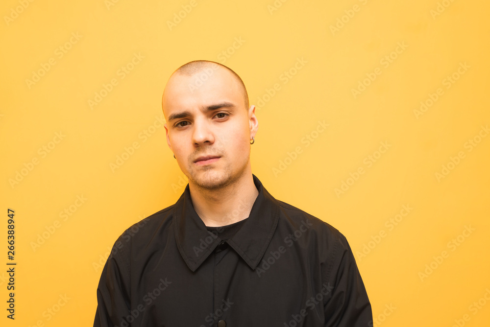 Stylish young man with a bald head wearing black jacket poses on a camera on a yellow background and looks seriously in the camera. Trendy authentic guy is isolated on yellow.