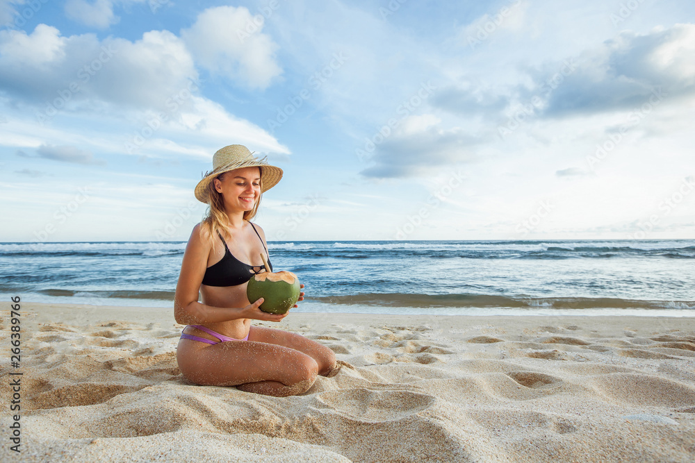 A girl in a hat and swimsuit sits on the beach holding a coconut. Copy space