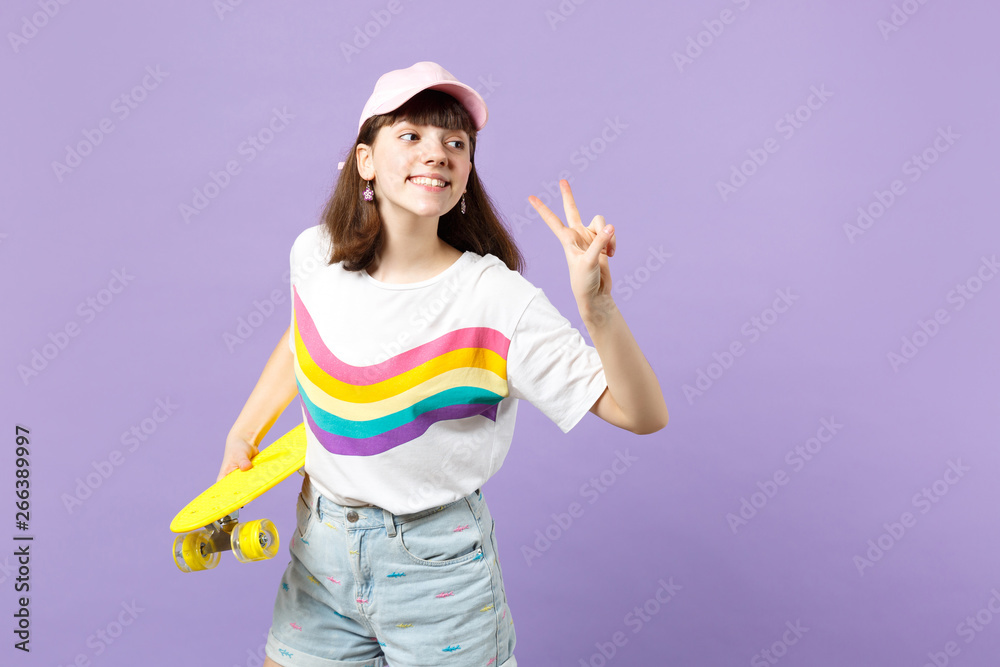 Charming teen girl in vivid clothes looking aside, holding yellow skateboard, showing victory sign isolated on violet pastel background. People sincere emotions, lifestyle concept. Mock up copy space. Charming teen girl in vivid clothes looking aside, holding yellow skateboard, showing victory sign isolated on violet pastel background. People sincere emotions, lifestyle concept. Mock up copy space.