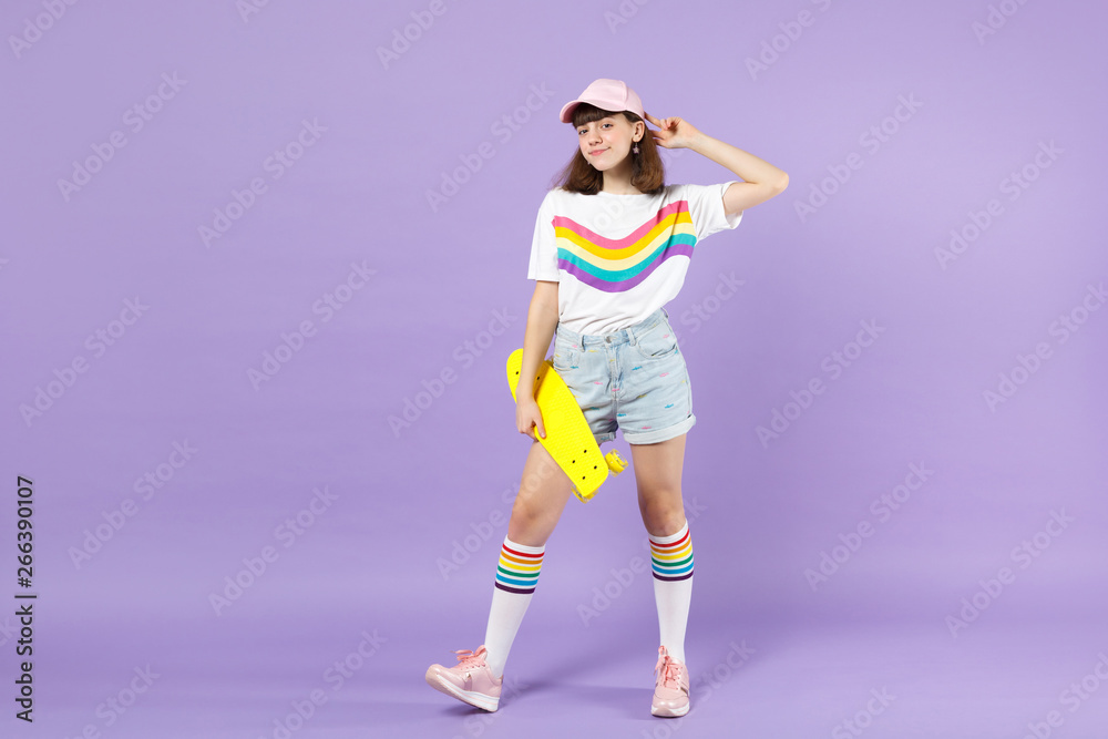 Portrait of pretty teen girl in vivid clothes standing, holding yellow skateboard isolated on violet pastel wall background in studio. People sincere emotions, lifestyle concept. Mock up copy space.
