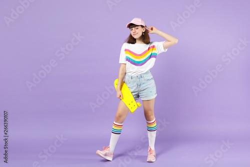 Portrait of pretty teen girl in vivid clothes standing, holding yellow skateboard isolated on violet pastel wall background in studio. People sincere emotions, lifestyle concept. Mock up copy space. © ViDi Studio
