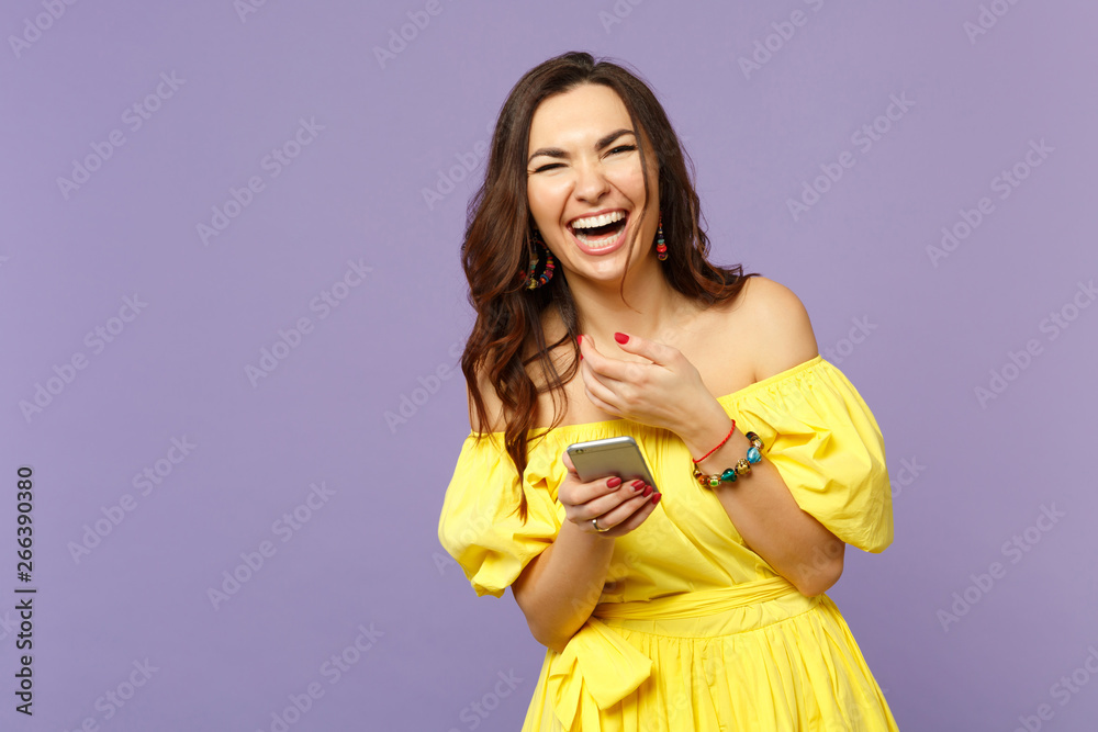 Laughing young woman in yellow summer dress using mobile phone, typing sms message isolated on pastel violet wall background in studio. People sincere emotions, lifestyle concept. Mock up copy space. Laughing young woman in yellow summer dress using mobile phone, typing sms message isolated on pastel violet wall background in studio. People sincere emotions, lifestyle concept. Mock up copy space.