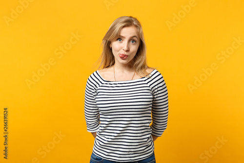 Portrait of funny young woman in striped clothes looking camera, showing tongue isolated on yellow orange wall background in studio. People sincere emotions, lifestyle concept. Mock up copy space.
