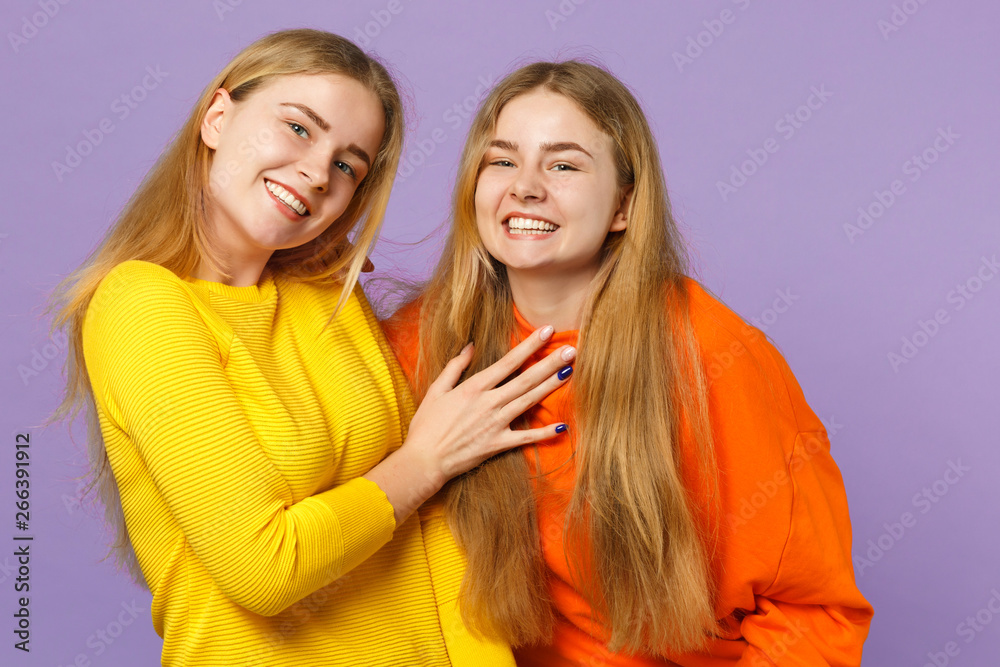 Two joyful cute young blonde twins sisters girls in vivid colorful clothes standing, looking camera isolated on pastel violet blue wall background. People family lifestyle concept. Mock up copy space.