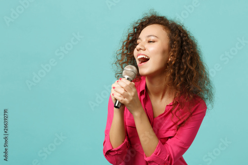 Portrait of beautiful african girl in casual clothes dancing sing song in microphone isolated on blue turquoise wall background in studio. People sincere emotion lifestyle concept. Mock up copy space.
