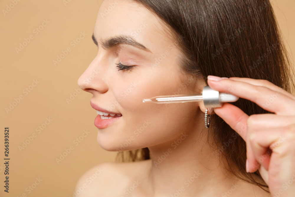 Close up brunette half naked woman 20s with perfect skin, nude make up apply oil from bottle on face isolated on beige pastel wall background, studio portrait. Healthcare cosmetic procedures concept. Close up brunette half naked woman 20s with perfect skin, nude make up apply oil from bottle on face isolated on beige pastel wall background, studio portrait. Healthcare cosmetic procedures concept.