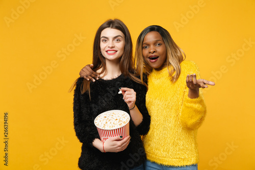 Φωτογραφία Two women friends european and african american in black yellow clothes hold bucket of popcorn isolated on bright orange wall background, studio portrait