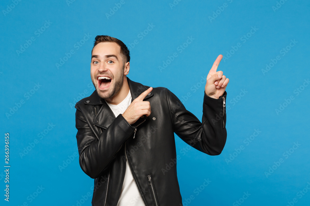 Handsome stylish young unshaven man in black jacket white t-shirt showing on workspace isolated on blue wall background studio portrait. People sincere emotions lifestyle concept. Mock up copy space. Handsome stylish young unshaven man in black jacket white t-shirt showing on workspace isolated on blue wall background studio portrait. People sincere emotions lifestyle concept. Mock up copy space.