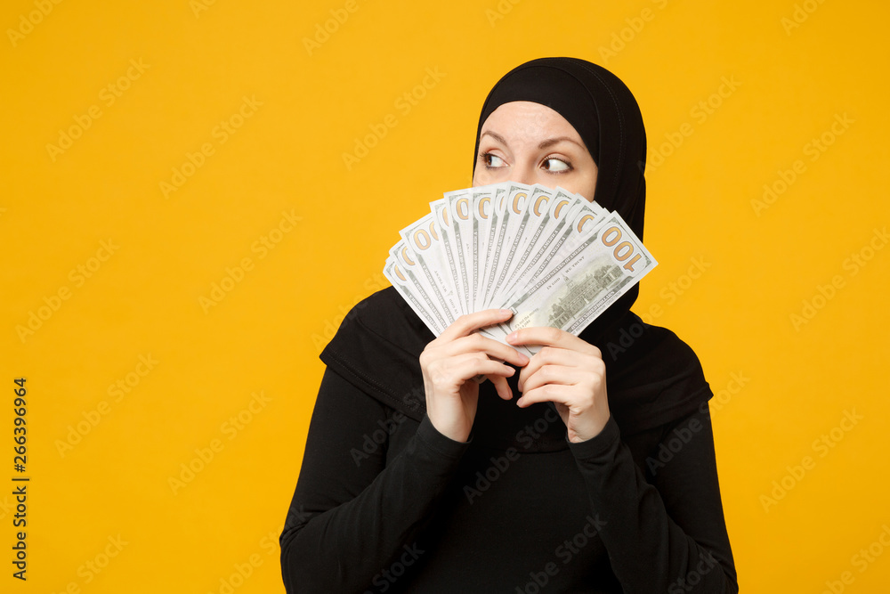 Young arabian muslim woman in hijab black clothes hold fan of cash money in dollar banknotes isolated on yellow wall background studio portrait. People religious lifestyle concept. Mock up copy space. Young arabian muslim woman in hijab black clothes hold fan of cash money in dollar banknotes isolated on yellow wall background studio portrait. People religious lifestyle concept. Mock up copy space.