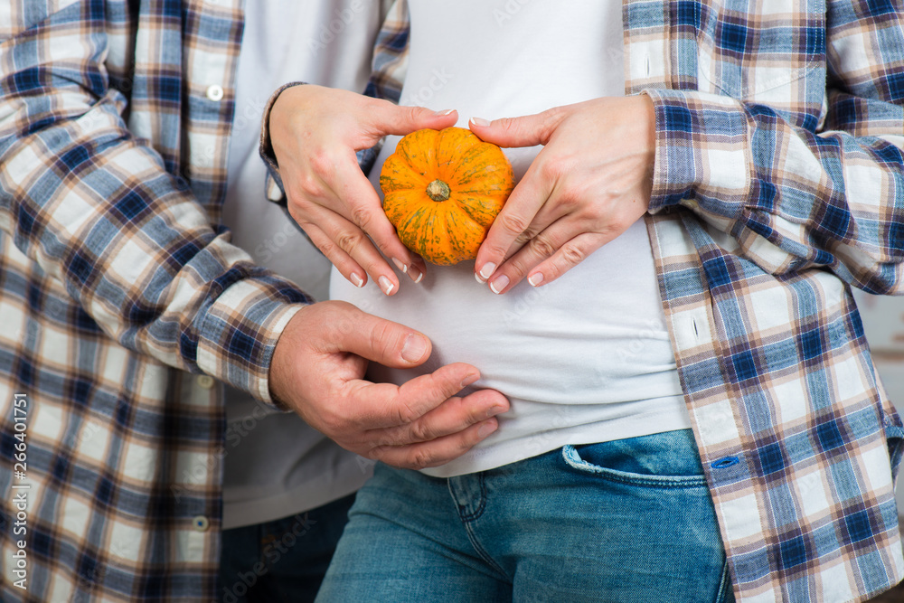 husband and wife in jeans and a plaid shirt holds little pumpkin in their hands. Couple posing a heart with their fingers on belly of expectant mother. husband and wife in jeans and a plaid shirt holds little pumpkin in their hands. Couple posing a heart with their fingers on belly of expectant mother.