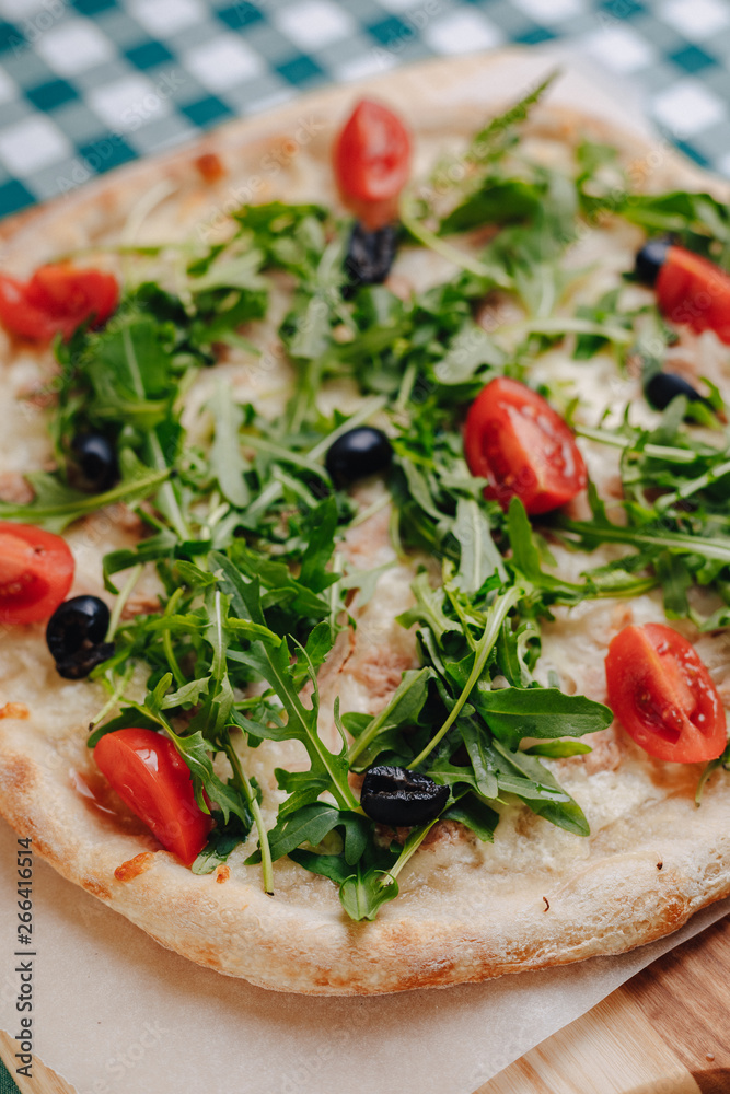 Neapolitan pizza with tuna, cheese, arugula, basil, tomatoes, olives, sprinkled with cheese on a wooden table on a tablecloth in a cell with a place for the text.