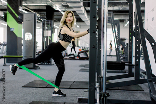 Sporty young blond woman does exercises on legs with sport fitness rubber bands. Photo of muscular woman in sportswear training in gym. Strength and motivation.