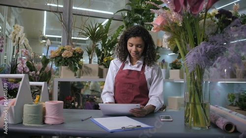 Mixed race female florist looking at tablet pc display accepting orders from customers on delivery of flowers. Modern floristical salon provides flower delivery service, employee networking at counter photo