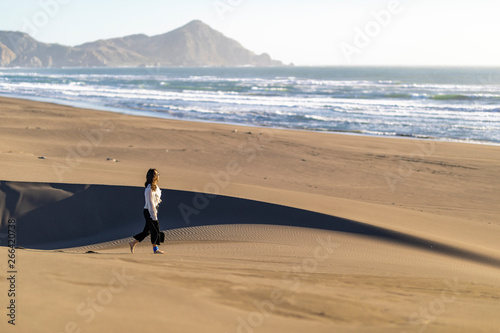 Beautiful young girl walking along the beach sand dunes step by step letting it footprints in the sand. An amazing wild beach during a sunny day at sunset time in Topocalma Beach, an awe coast scenery