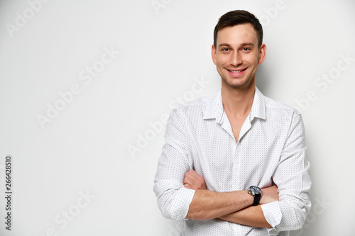 Young man with a wide kind smile stands at the white wall with arms crossed near free text space