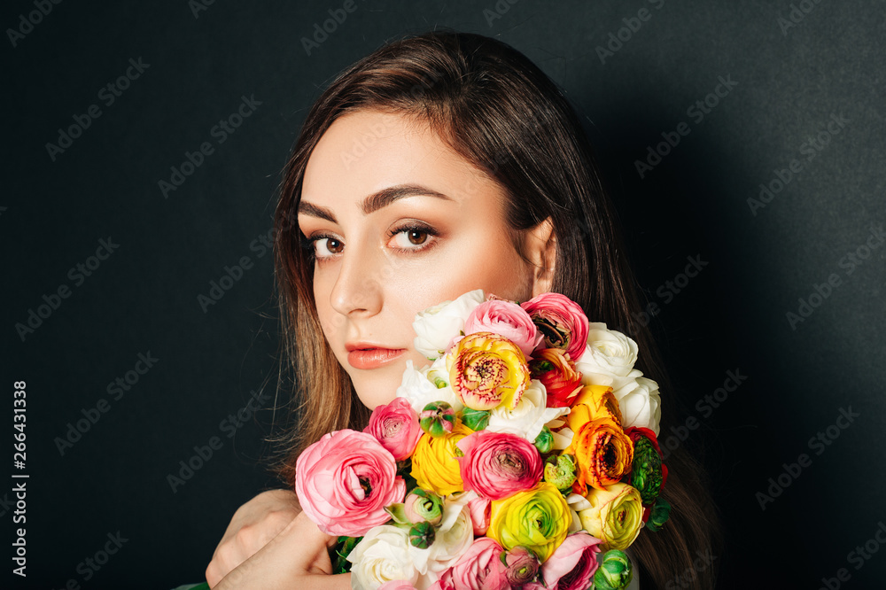 Studio close up portrait of beautiful young woman with long shiny hair, professional makeup, wearing white dress, holding colorful buttercup flowers Studio close up portrait of beautiful young woman with long shiny hair, professional makeup, wearing white dress, holding colorful buttercup flowers
