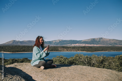 A young brunette woman is sitting on the stone in the middle of the nature. She is looking her phone while writing something. She is enjoying the nature in a beautiful sunny day.