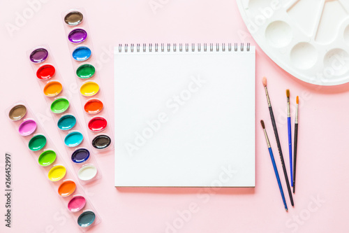 Creative flat lay of watercolor palettes, paint brushes, white paper. Artist workplace on a pink pastel background.