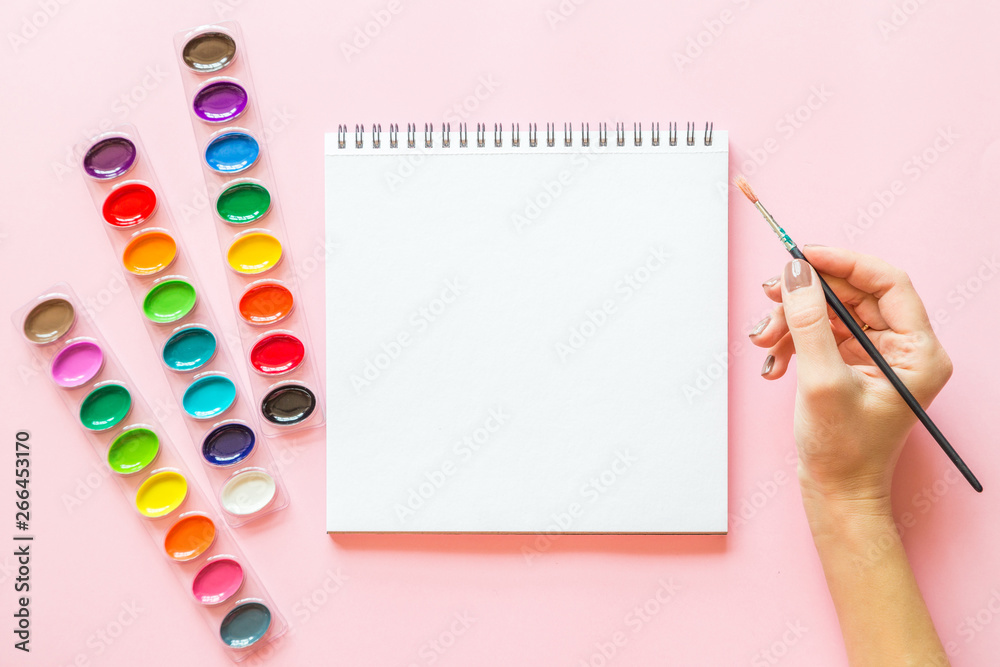 Creative flat lay of watercolor palettes, notebook, female hand holding paint brush. Artist workplace on a pink pastel background.