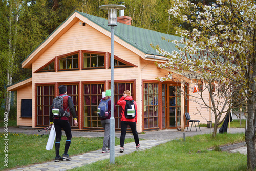 Three athletes wearing running backpacks man and two women participants of orienteering or rogaining sport competition walking on pathway to wooden house information centre.