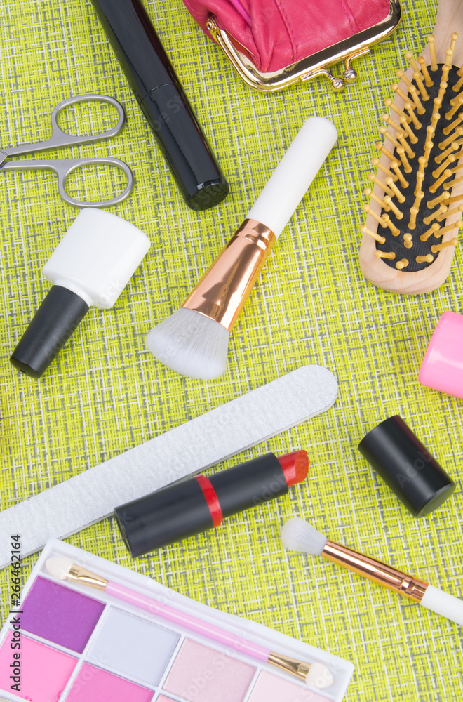 on a green background, tools for manicure and makeup, nail polishes, scissors, red lipstick, powder brushes, colorful eye shadow, mascara and hairbrush