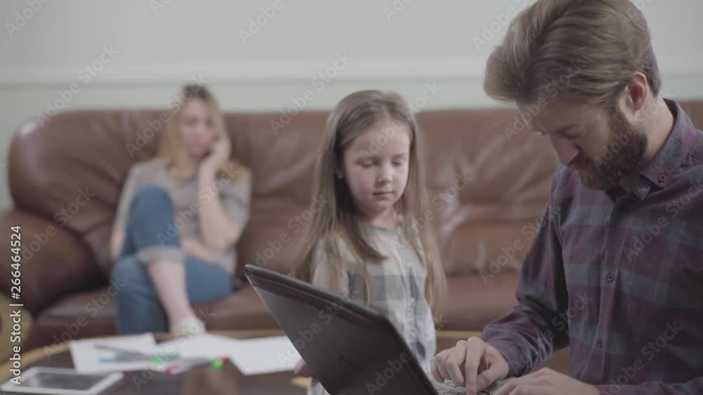 Portrait of the bearded man working with laptop close up. Little girl unsuccessfully trying to give the man the apple while mother sitting on the sofa on the background. Young man addicted to gadgets Portrait of the bearded man working with laptop close up. Little girl unsuccessfully trying to give the man the apple while mother sitting on the sofa on the background. Young man addicted to gadgets