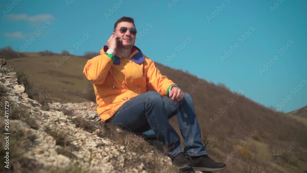 A young man in a yellow jacket, blue jeans and glasses sits in the mountains and enjoys the scenery and talking on the phone, laughing. In the background are hills and sky. Mountain landscape. A young man in a yellow jacket, blue jeans and glasses sits in the mountains and enjoys the scenery and talking on the phone, laughing. In the background are hills and sky. Mountain landscape.