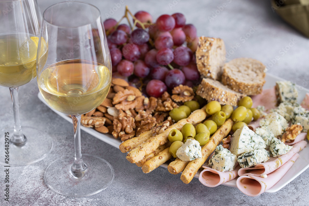 Two glasses of white wine and plate with different snacks. Blue cheese, olives, baguette slices, grissini, ham, grapes and nuts. Wine snacks set background. Two glasses of white wine and plate with different snacks. Blue cheese, olives, baguette slices, grissini, ham, grapes and nuts. Wine snacks set background.
