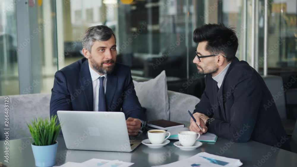 Colleagues handsome middle-aged men in suits are talking in cafe during business lunch using laptop sitting at table discussing work. People and talks concept.