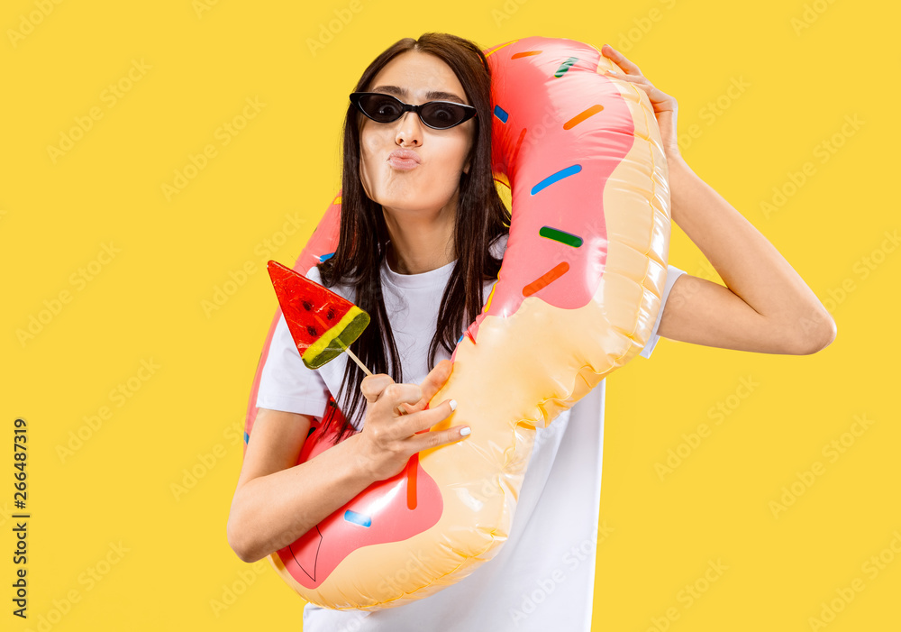 Beautiful female half-length portrait isolated on yellow studio background. Young smiling woman in black sunglasses with candy. Facial expression, summer, weekend, resort concept. Trendy colors.