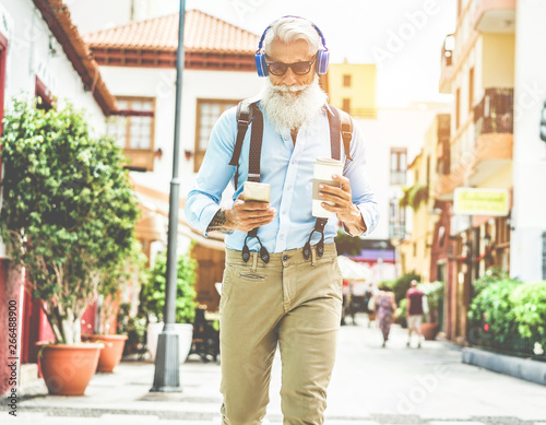 Trendy senior man using music smartphone app and drinking coffee in downtown center outdoor - Mature fashion male having fun with new trends technology - Tech and joyful elderly lifestyle concept