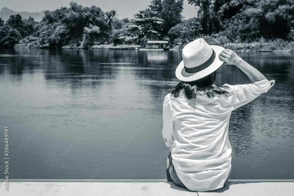 Lonely Asian woman wear weave hat and white shirt sitting on wooden terrace and looking forward to the river.
