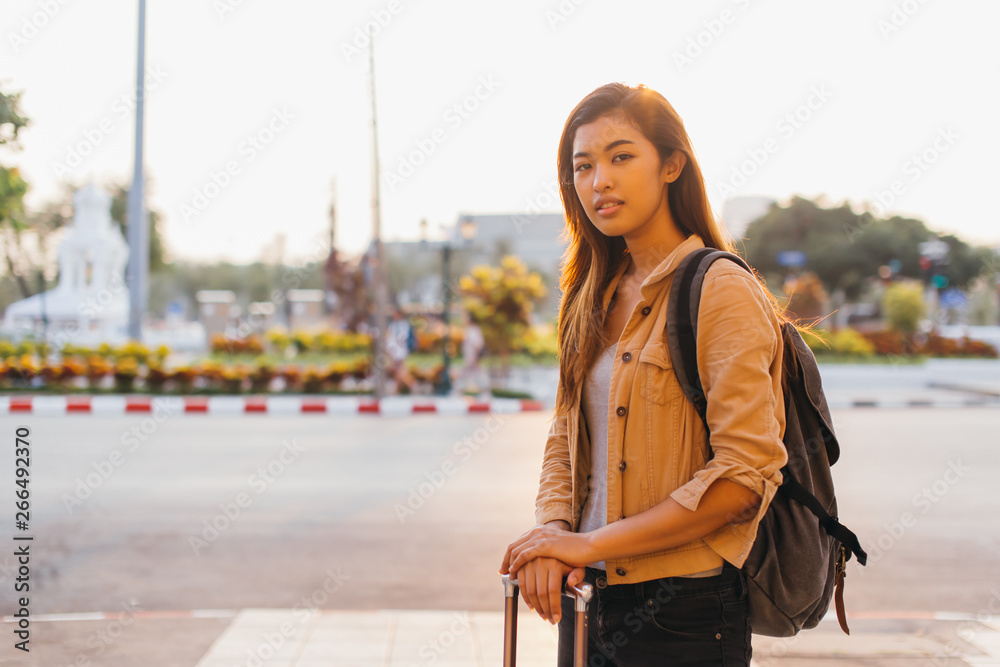 Smiling Asian woman with backpack and luggage standing on street in sunlight looking at camera, Bangkok, Thailand
