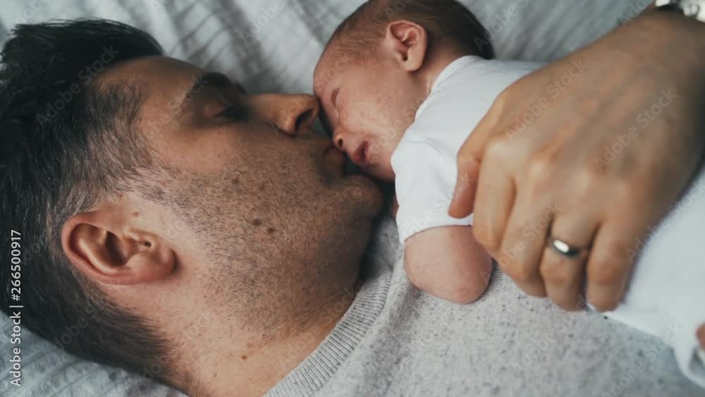 Touching, tender scene as infant sleeps on the chest of his father. Daddy holding his baby close as they both prepare for a nap. Dad kissing baby snuggled to his chest. Touching, tender scene as infant sleeps on the chest of his father. Daddy holding his baby close as they both prepare for a nap. Dad kissing baby snuggled to his chest.