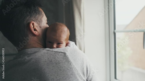 Dad holding baby near a bright window. Father comforts and cuddles infant while standing. Parenthood, fatherhood and love concept. Man with baby against his chest, head on shoulder. Slow motion. photo