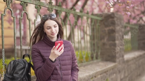 Young Caucasina Female Standing on Street Unders Spring Tree Looking at Mobile Phone and Smiles. Light Camera Movement and Blurred Background photo