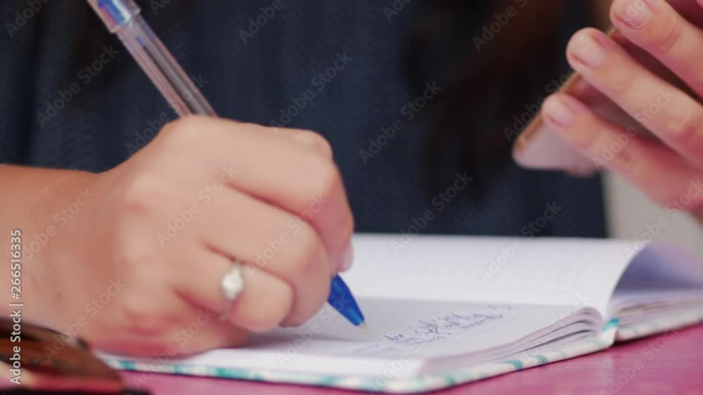 Young woman writing in a notebook with a blue pen noted from a cell phone Young woman writing in a notebook with a blue pen noted from a cell phone